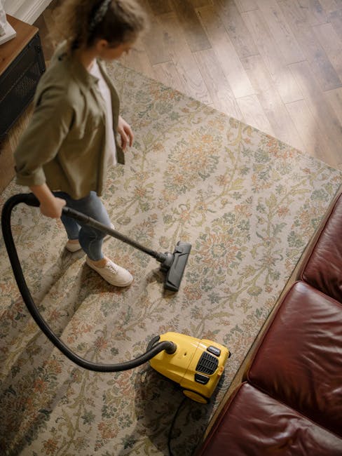 A woman is engaged in surface cleaning on a patterned, light-colored area rug in a living room, using a yellow vacuum cleaner. The room features wooden flooring adjacent to the rug, with natural light illuminating the space. Part of a red leather sofa is visible in the corner, and the woman is dressed casually in a light beige jacket, jeans, and sneakers. This scene demonstrates a professional deep cleaning process, emphasizing hygiene and maintenance, consistent with services offered by Carpet Cleaners Balham as part of their domestic cleaning and carpet care. The overall cleanliness appears high, with well-maintained surfaces and no visible dust or debris.