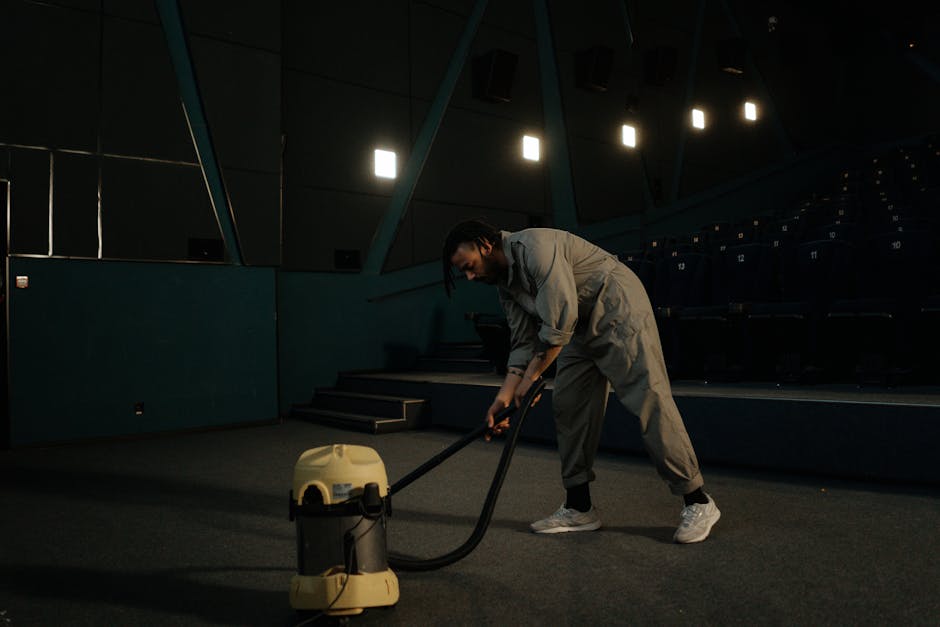 A person engaged in deep cleaning inside a dimly lit auditorium or theater, operating a large carpet or floor cleaning machine with a yellow body and black hose. The individual is dressed in grey overalls and sneakers, leaning forward to maneuver the equipment across a dark, carpeted surface. The background features empty tiered seating with dark upholstery and modern geometric wall panels illuminated by overhead lights, creating a professional cleaning environment. This scene represents surface cleaning and maintenance, highlighting the expertise of [COMPANY_NAME] in commercial carpet cleaning, as detailed in the Balham High Road carpet cleaning guide.
