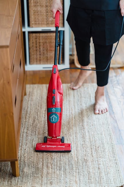 A woman is engaged in surface cleaning on a patterned, light-colored area rug in a living room, using a yellow vacuum cleaner. The room features wooden flooring adjacent to the rug, with natural light illuminating the space. Part of a red leather sofa is visible in the corner, and the woman is dressed casually in a light beige jacket, jeans, and sneakers. This scene demonstrates a professional deep cleaning process, emphasizing hygiene and maintenance, consistent with services offered by Carpet Cleaners Balham as part of their domestic cleaning and carpet care. The overall cleanliness appears high, with well-maintained surfaces and no visible dust or debris.